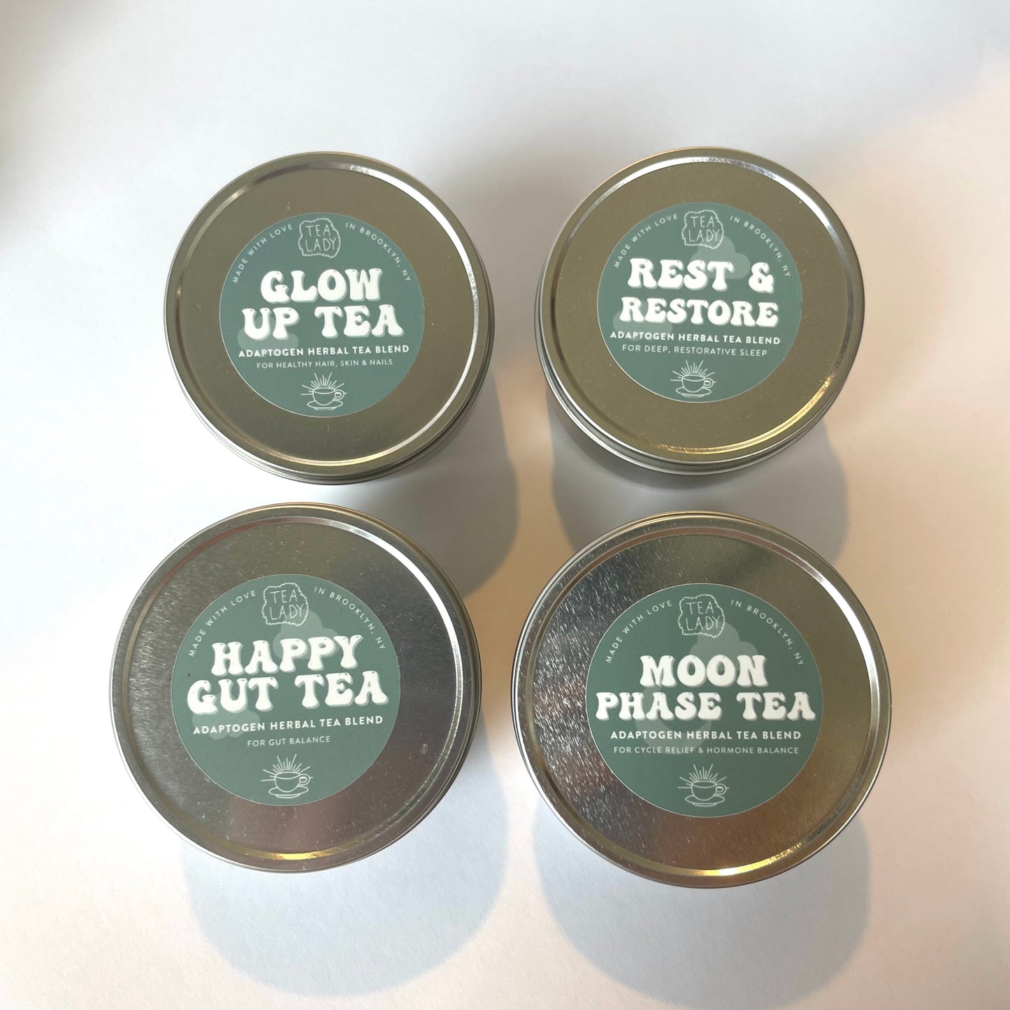 4 tins with green stickers labeled: glow up tea, rest & restore, Happy gut tea, moon phase tea