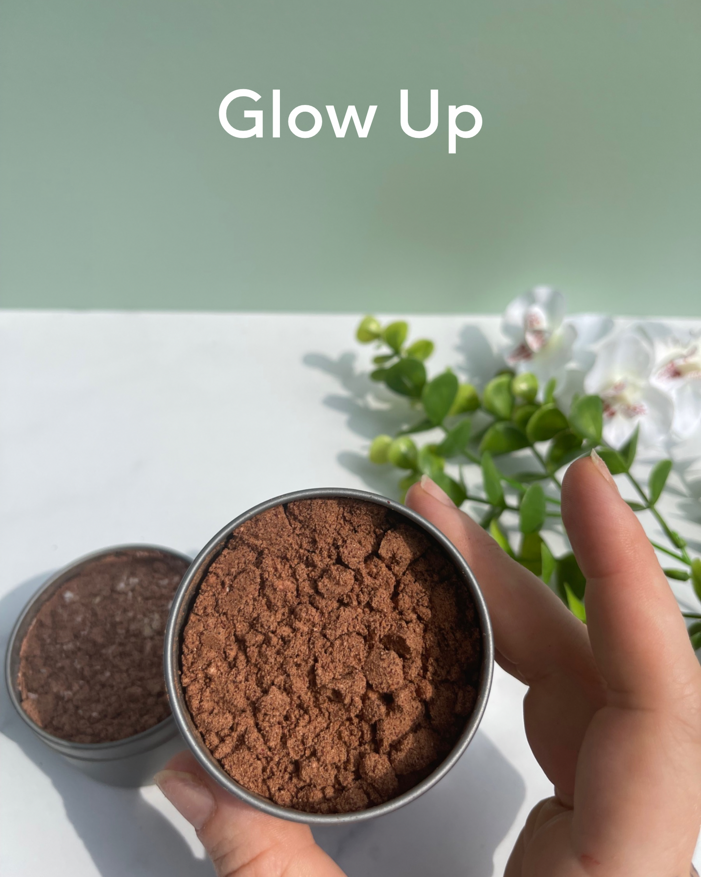 Reads: Glow UP. Foreground : hand holding metal tin containing herbal blend. Background: 2 metal tins containing herb blends sitting on marble counter next to flowers