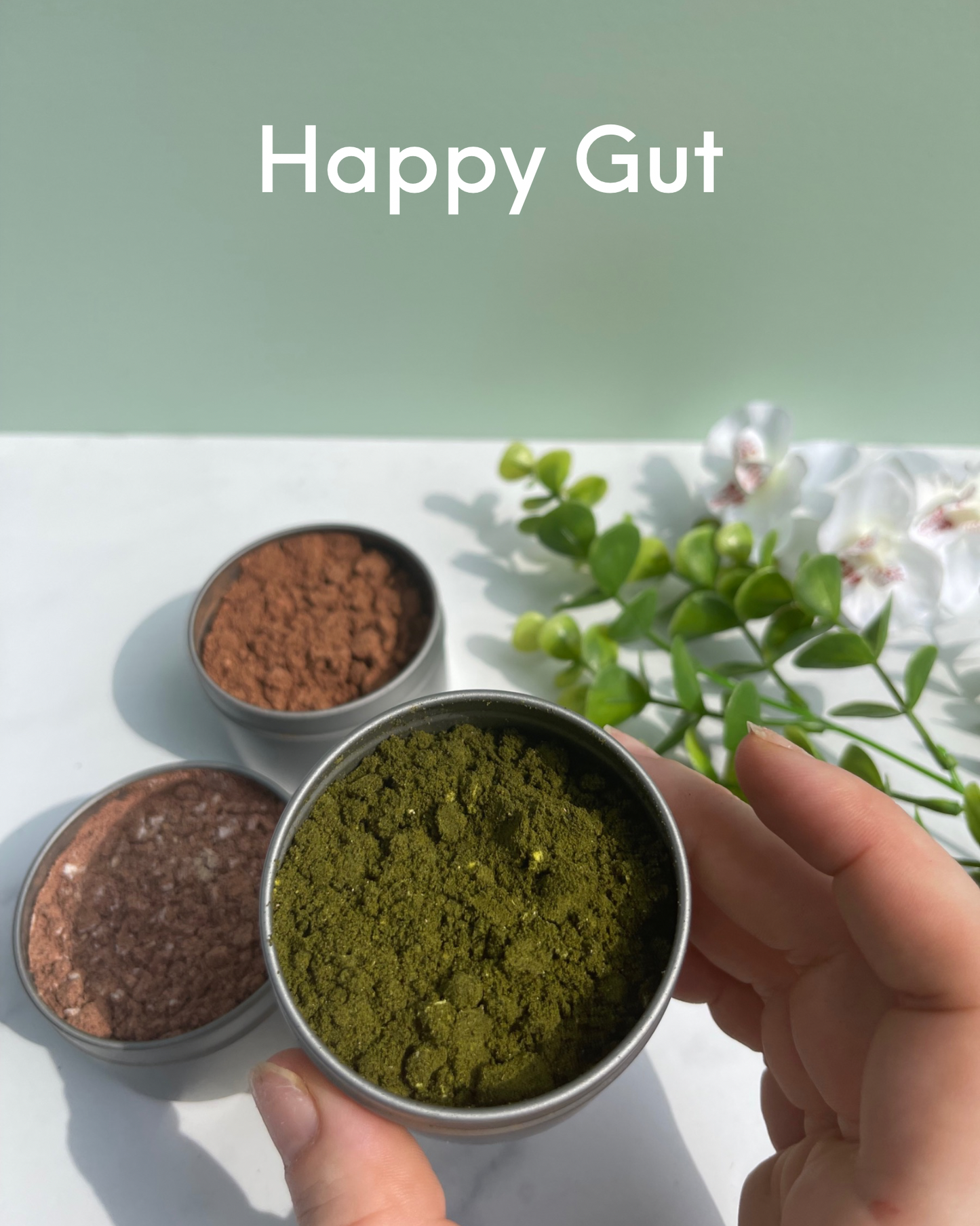 Reads: Happy Gut. Foreground : hand holding metal tin containing herbal blend. Background: 2 metal tins containing herb blends sitting on marble counter next to flowers
