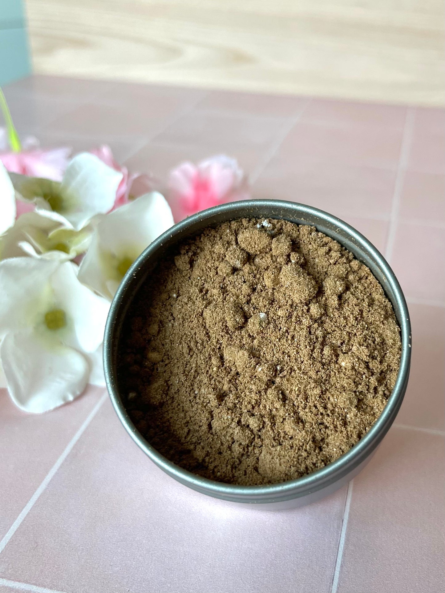 Metal tin containing herbal powder blend sitting on tile counter next to flowers