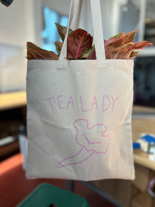 Off white tote bags with Screen printed TEA LADY and trumpet mushroom. Plant leaves are poking out the top of the tote bag