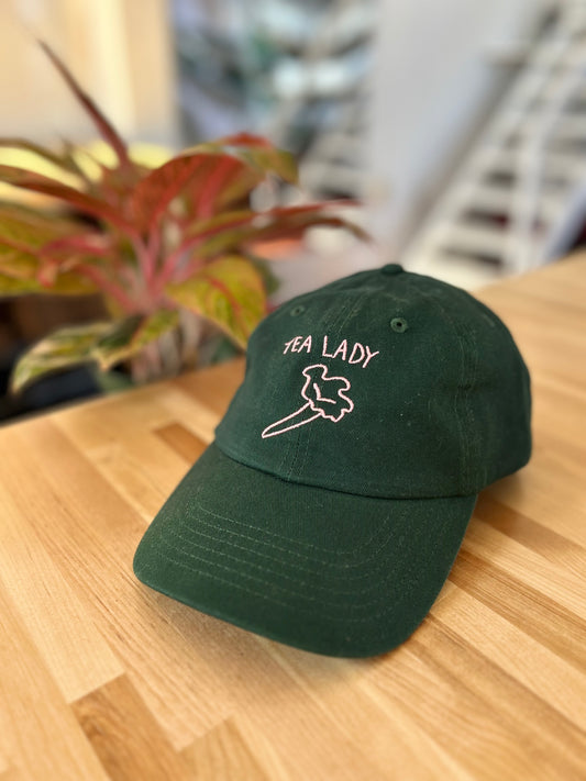 Forest green cap with pink stitching of trumpet mushroom. Pink stitching reads TEA LADY. Cap is sitting on wood table next to plant